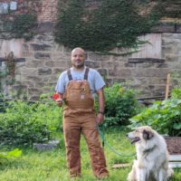 Timothy, founder, in coveralls holding a flower and smiling as his dog stares back at him, smiling in Unity Garden
