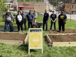 Eight humans stand behind two freshly built planter boxes with a wheel barrow and shovels in between. A person's head peeks above an a frame sign that states "Unity Garden, where community grows". The lot sits in front of a Family Dollar store. 